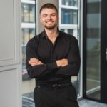 Mike Grammel, Gründer und Geschäftsführer von BetonGold Immobilien, steht lächelnd mit verschränkten Armen in einem modernen Bürogebäude.