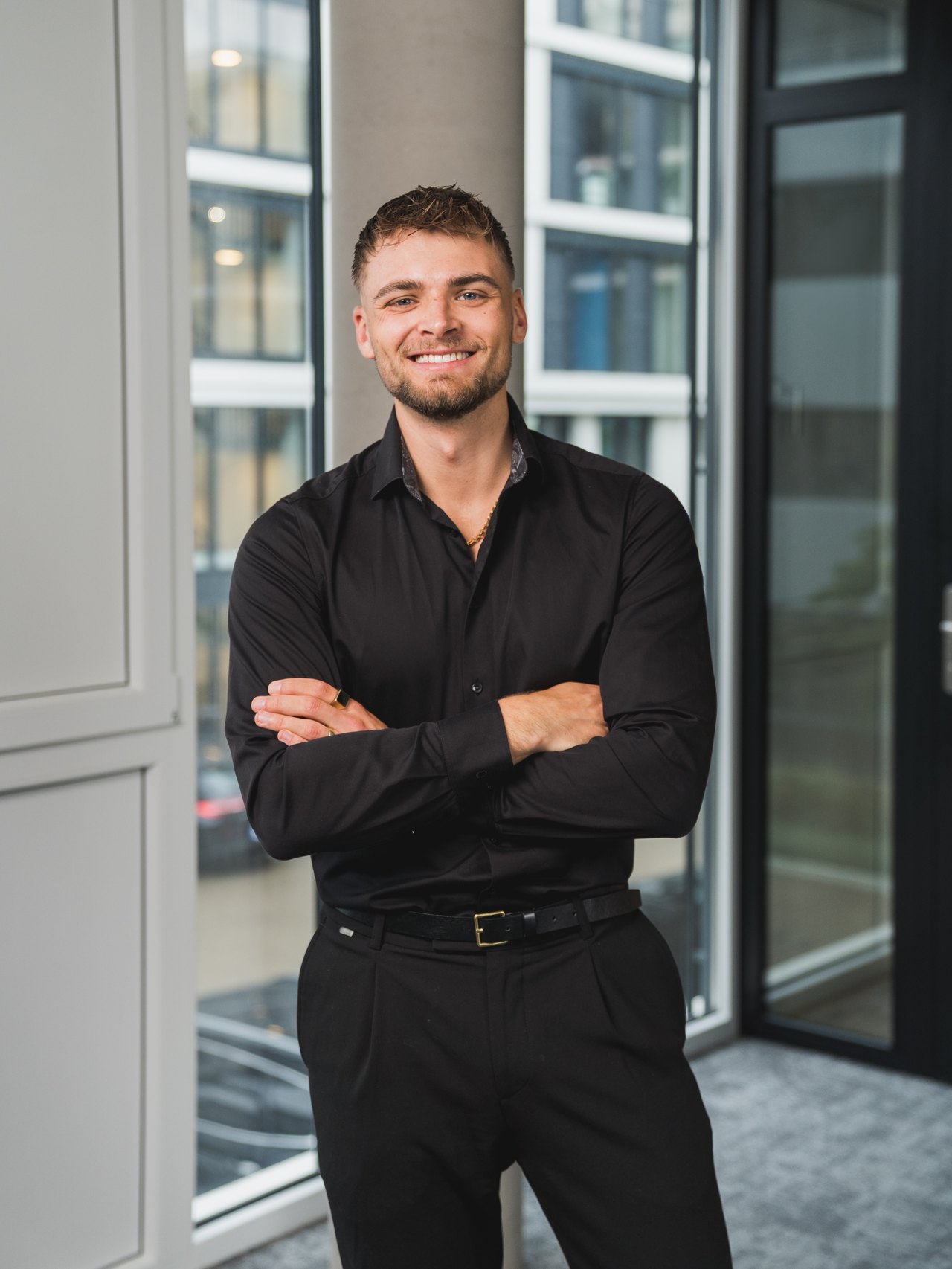 Mike Grammel, Gründer und Geschäftsführer von BetonGold Immobilien, steht lächelnd mit verschränkten Armen in einem modernen Bürogebäude.
