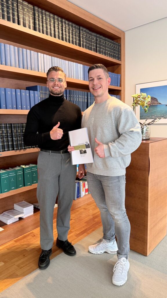 Mike Grammel und Daniel Kubik zeigen Daumen hoch und halten eine Broschüre in einem modernen Büro mit Bücherregalen