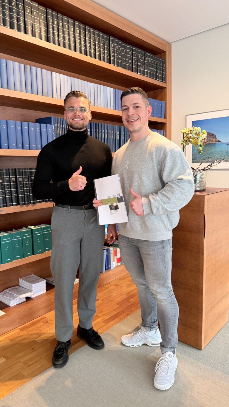 Mike Grammel und Daniel Kubik zeigen Daumen hoch und halten eine Broschüre in einem modernen Büro mit Bücherregalen