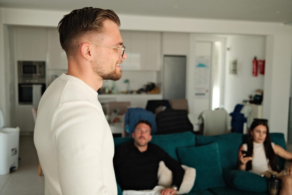 Mike Grammel steht im Vordergrund eines hellen Wohnraums, während zwei Teammitglieder im Hintergrund auf einem Sofa sitzen.