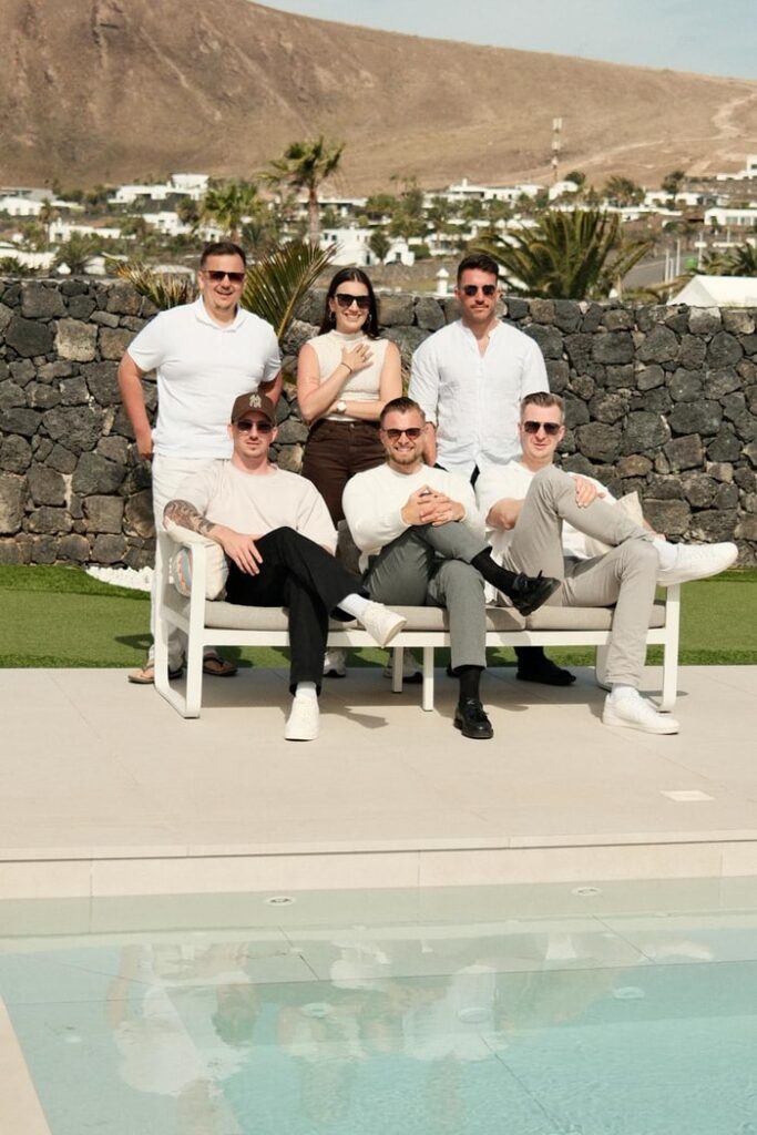 Das Team von BetonGold Immobilien posiert in heller Kleidung vor einer Steinmauer, mit Pool, Palmen und bergiger Landschaft im Hintergrund.
