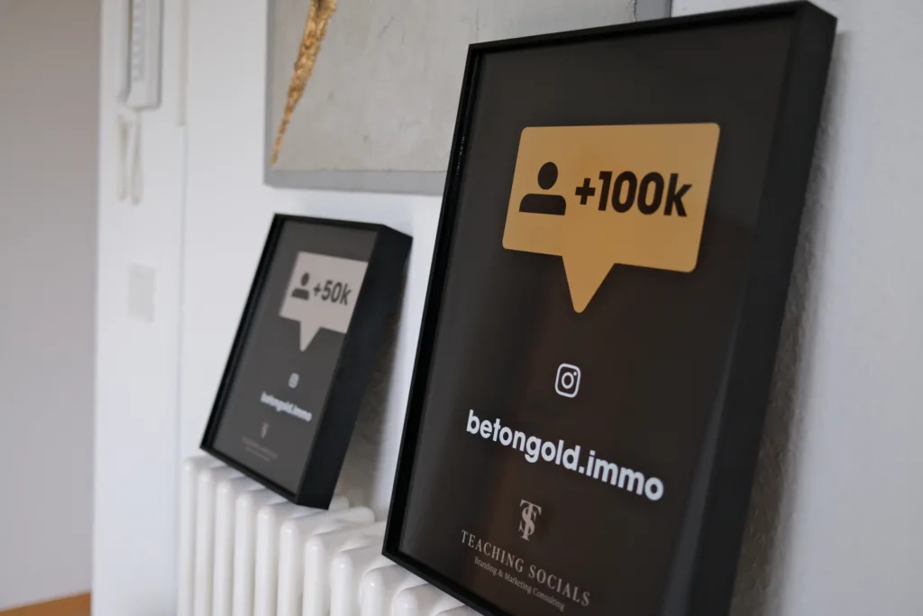 Zwei gerahmte Schilder an einer Wand, die Social Media Erfolge mit Abonnentenzahlen und dem Logo von BetonGold Immobilien präsentieren