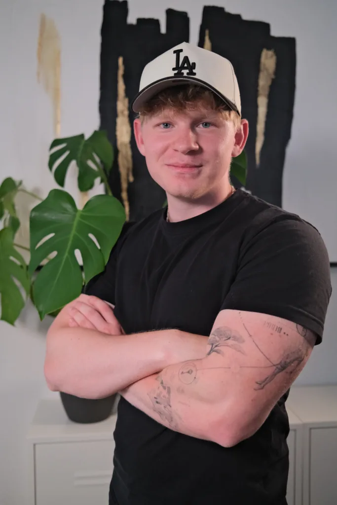 Alexander Darras steht mit verschränkten Armen vor einem Kunstwerk. Im Hintergrund ist eine große Pflanze sichtbar